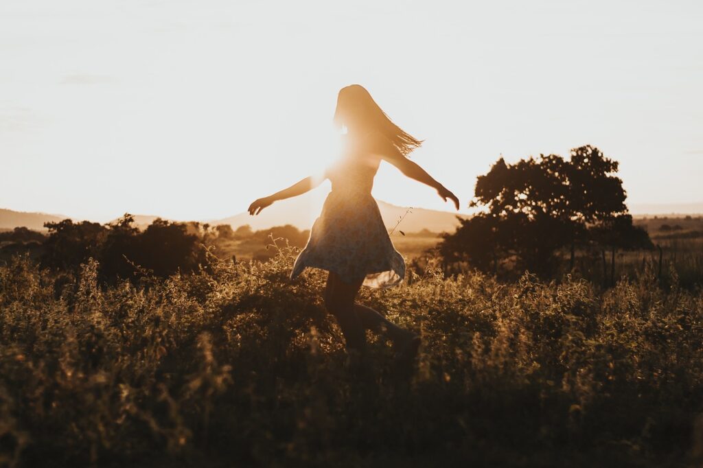 À contre jour, dans un champs au soleil couchant, une femme en robe les cheveux long et détachés danse, les bras semblant s'élever autour d'elle.