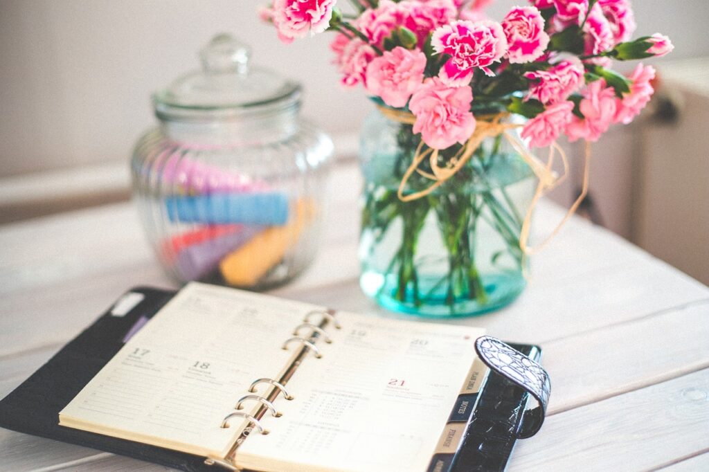 Pour illustrer la prise de rendez-vous, agenda ouvert devant un vase de fleur roses. En arrière plan un pot en verre contenant des feutres de couleurs.